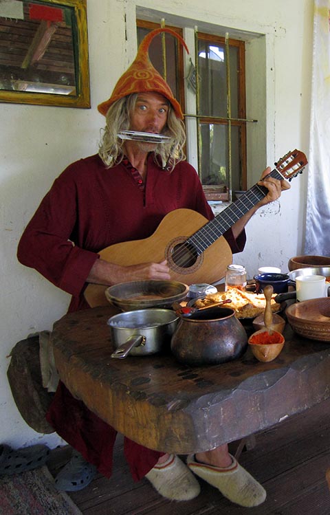 Istvan Sky in Narrenkappe und bordeauxfarbener Kurta mit Gitarre und Mundharmonika am Eichentisch auf der Veranda seines Hauses Napfényszentély in Szentbékkállá, Ungarn.
