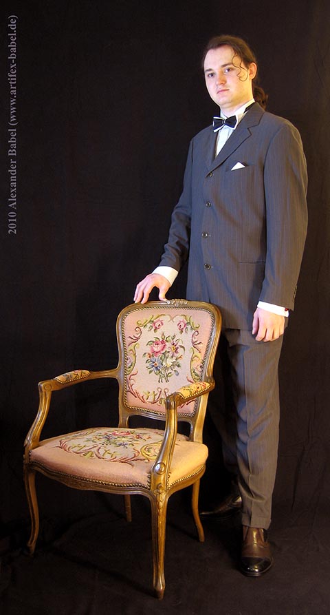 Alexander Babel in Großaufnahme, Ganzkörper, frontal. Im Anzug mit Fliege. Daneben ein antiker Stuhl.