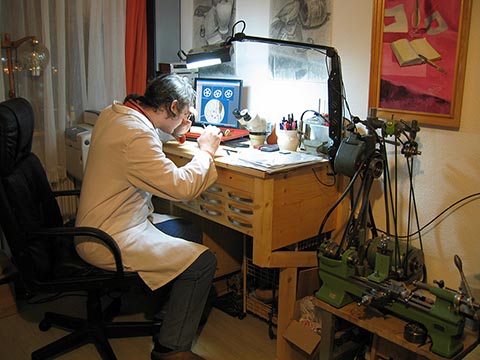 Alexander Babel arbeitet am Uhrmachertisch, daneben auf der Halterung die Drehmaschine Schaublin 70, an den Wänden eigene Zeichnungen.